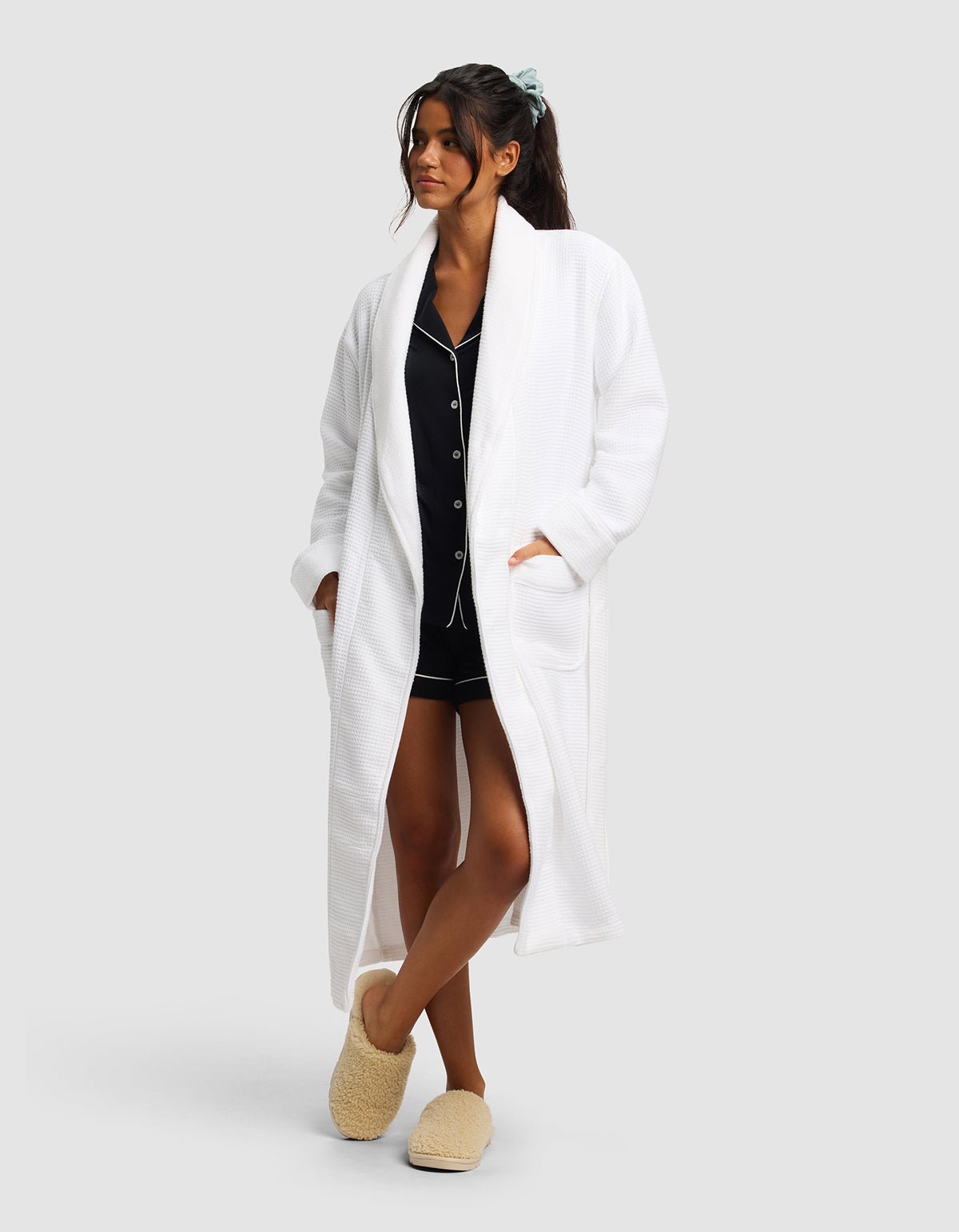 A woman stands against a plain light background wearing Cozy Earth's Waffle Bath Robe over black pajamas, with her hands in the robe's pockets. She pairs the look with fluffy slippers and a blue scrunchie in her hair.|Color:White