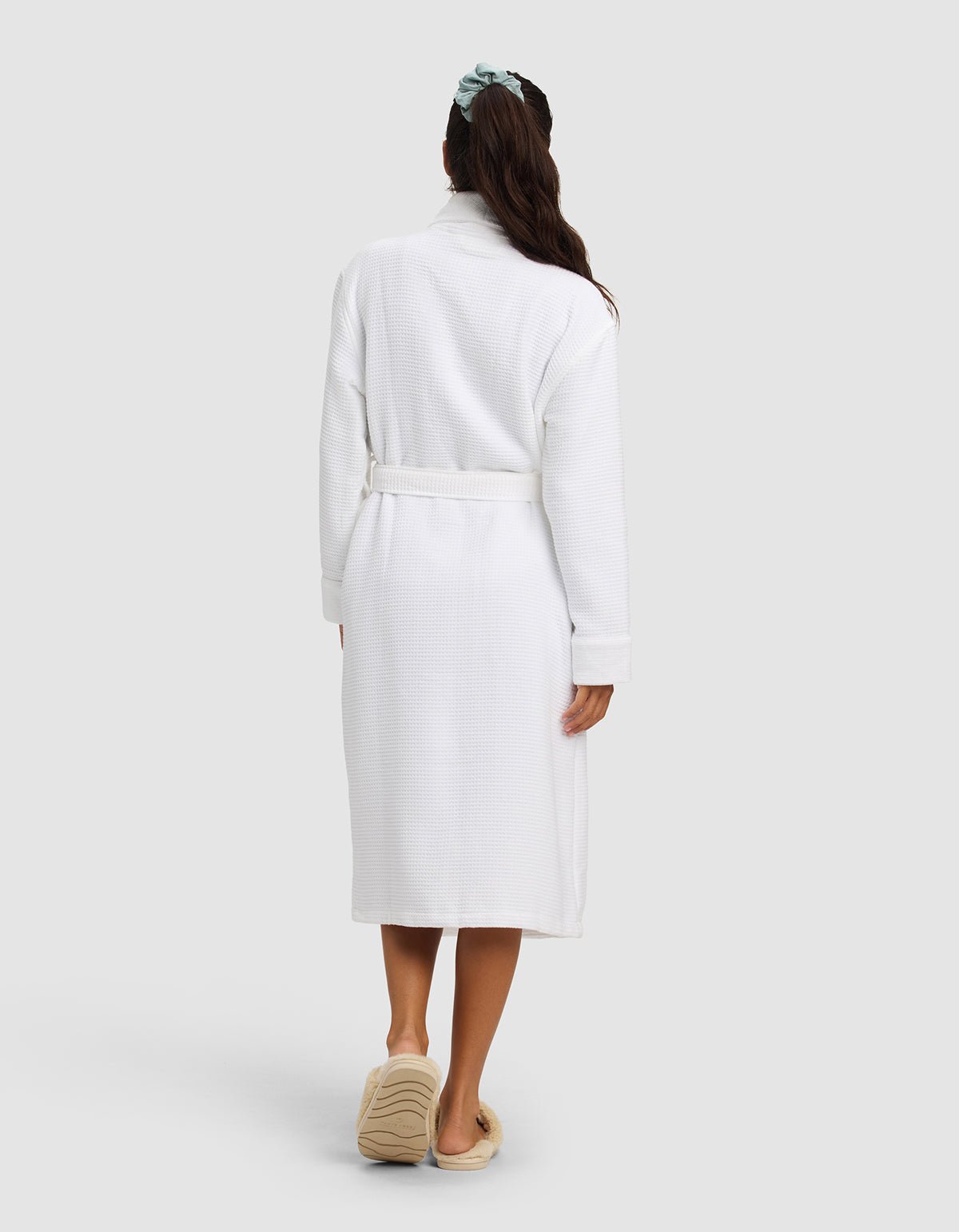 A woman with long dark hair in a green scrunchie faces away, wearing the Cozy Earth Waffle Bath Robe in white and tan slippers, standing against a plain light gray background.|Color:White