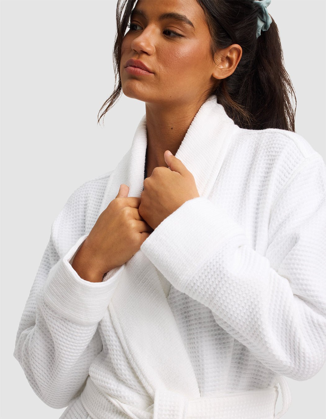 A woman with a blue scrunchie wears the Cozy Earth Waffle Bath Robe, holding the collar with both hands and gazing to the side against a plain light background.|Color:White