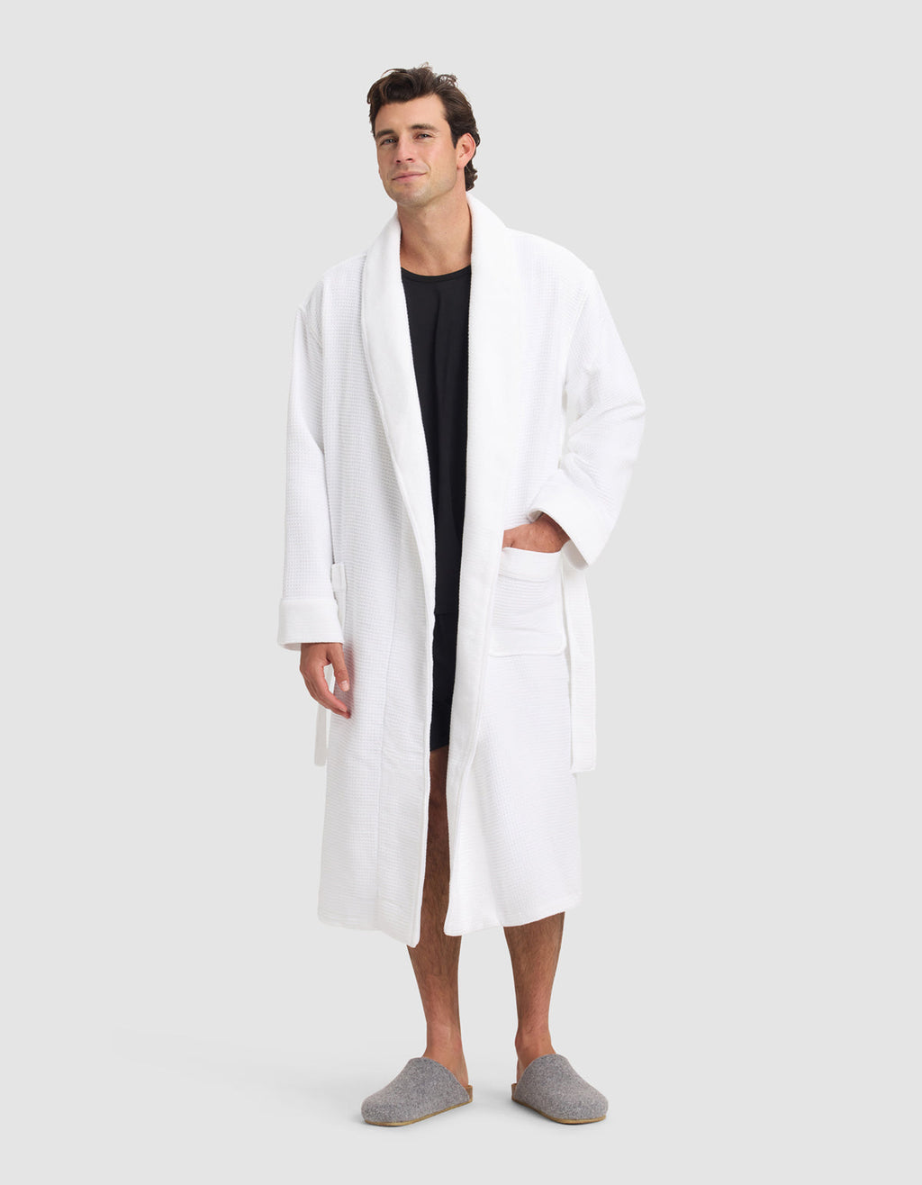 A man stands facing forward against a plain light grey background, wearing the Cozy Earth Waffle Bath Robe over a black shirt with grey slippers. One hand rests in the robe’s pocket.|Color:White