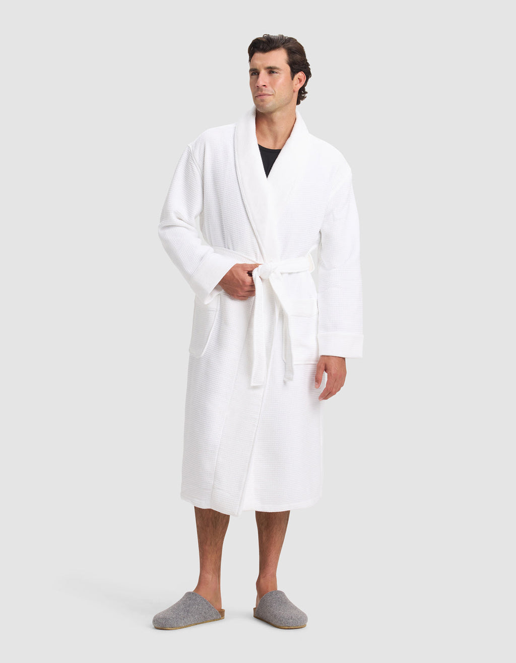 A man wears the Cozy Earth Waffle Bath Robe with a tied belt over a dark shirt, paired with gray slippers, standing against a plain light gray background.|Color:White