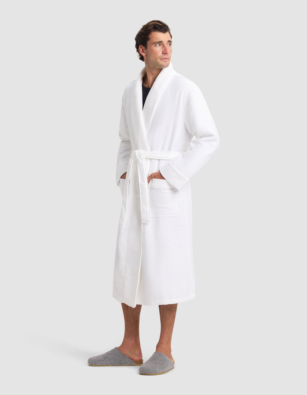 A man stands against a light gray background, wearing the Cozy Earth Waffle Bath Robe with a belted waist and pockets, along with gray slippers. He looks slightly to the side.|Color:White
