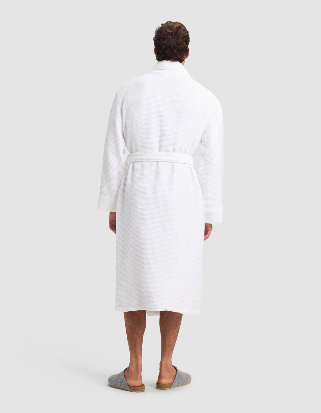 A person faces away, wearing the Cozy Earth Waffle Bath Robe—a white, knee-length robe with a belt—paired with gray slippers against a plain light background.|Color:White