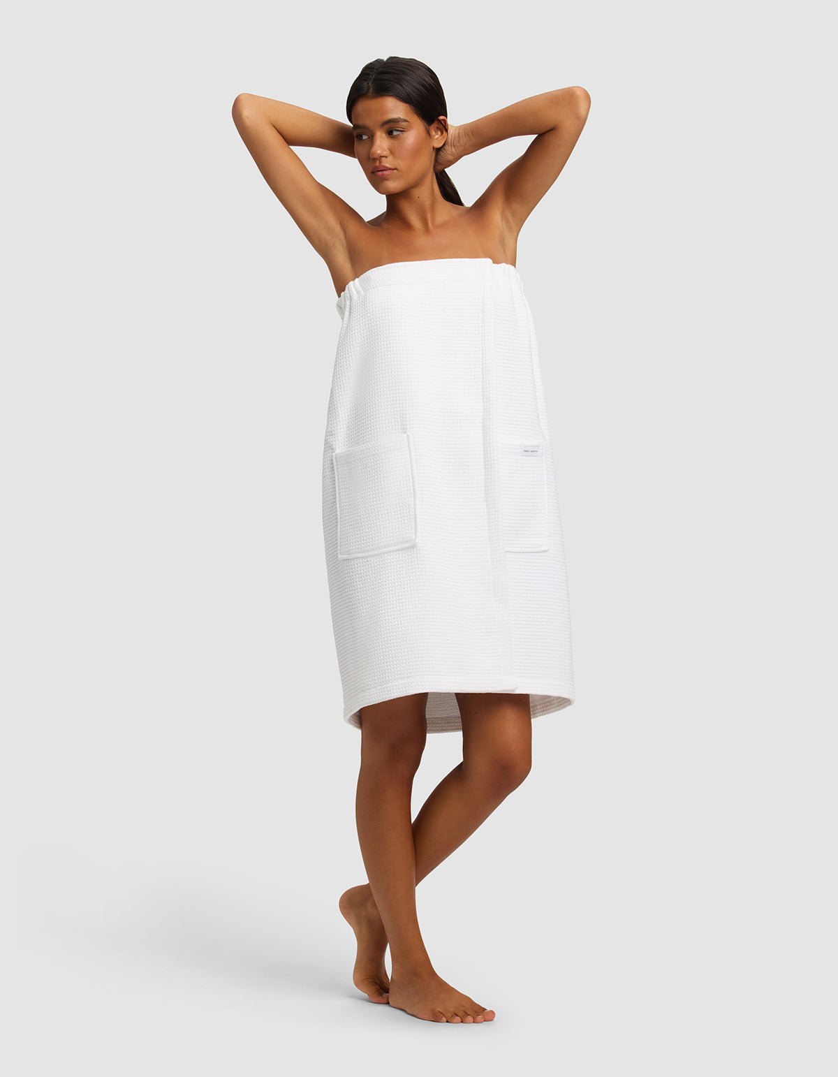 A woman stands barefoot with her arms raised, wearing the Cozy Earth Waffle Bath Wrap with large front pockets against a plain light gray background.|Color:White