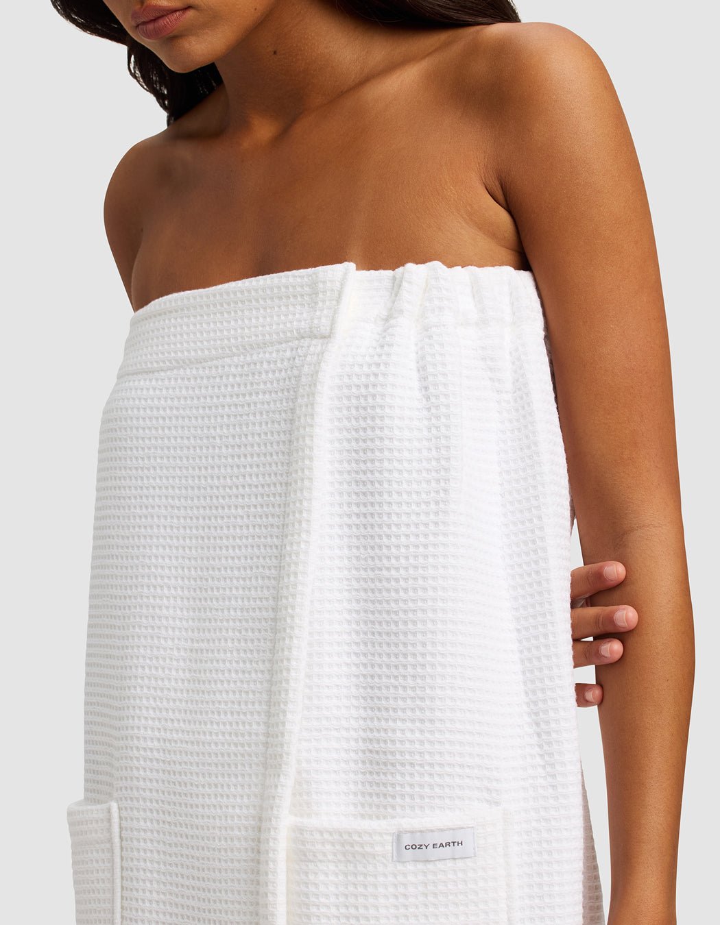 A woman with long dark hair is wrapped in the Cozy Earth Waffle Bath Wrap, a white, textured towel with pockets. She stands against a plain light gray background, shown from shoulders to mid-thigh.|Color:White