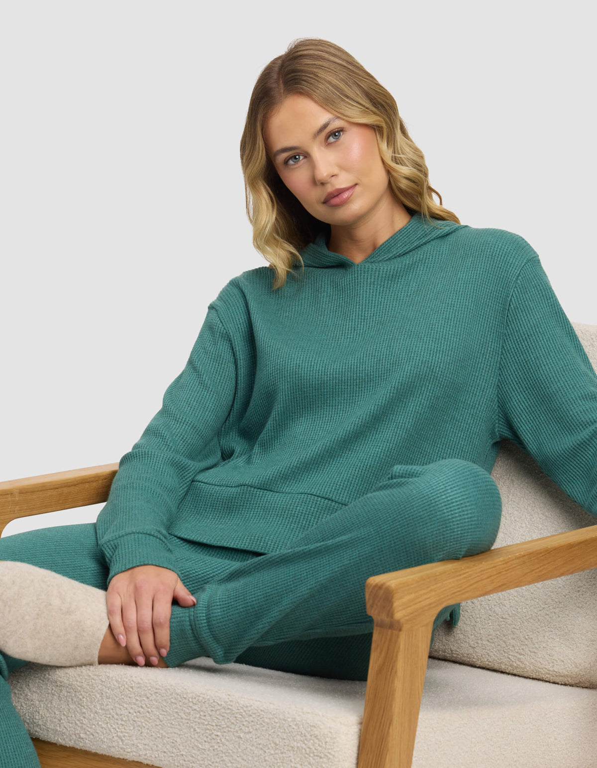 A blonde woman sits relaxed on a beige armchair, wearing the Cozy Earth Women's Blake Waffle Hoodie in teal, with one arm resting on the chair and her legs crossed.|Color:Sea Green