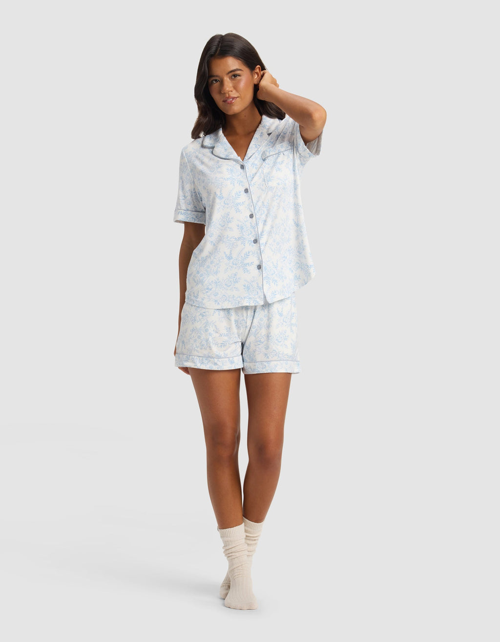 A woman stands in a relaxed pose wearing the Cozy Earth Women's Bamboo Stretch-Knit Short Sleeve Pajama Set with matching shorts and cream socks against a plain light background.|Color:Sky Toile