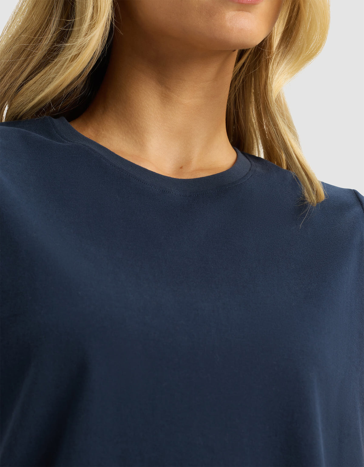 Close-up of a woman wearing the Cozy Earth Women's All Day Sleeveless Tee in navy blue, showing her neck, collar, and part of her blonde hair against a neutral light gray background.|Color:Eclipse