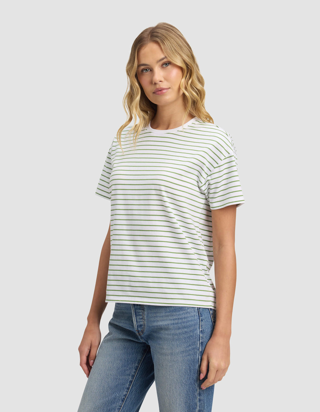 A woman with wavy blonde hair models the Cozy Earth Women's All Day Tee, a white shirt with thin green stripes, paired with blue jeans against a plain light gray background, glancing slightly to the side.|Color:Clover Stripe