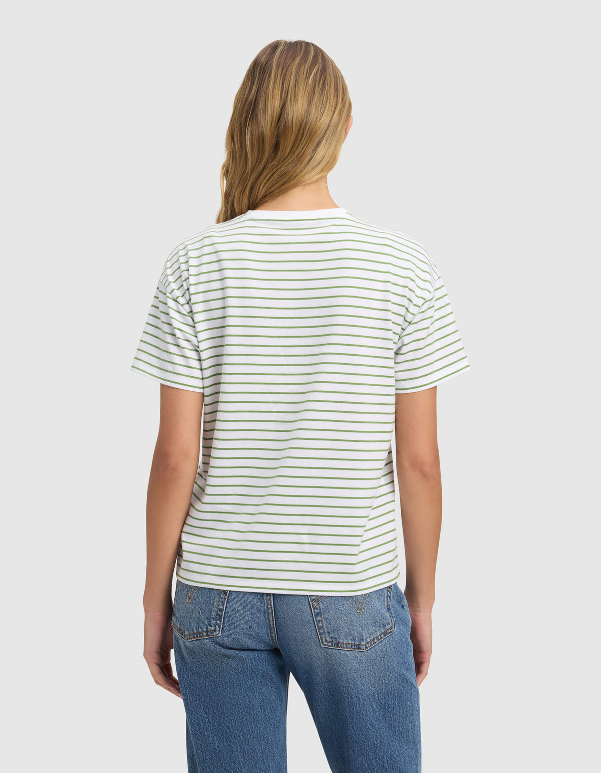 A woman with long, light brown hair faces away wearing the Cozy Earth Women's All Day Tee—white with thin horizontal black stripes—and blue jeans, set against a plain light gray background.|Color:Clover Stripe