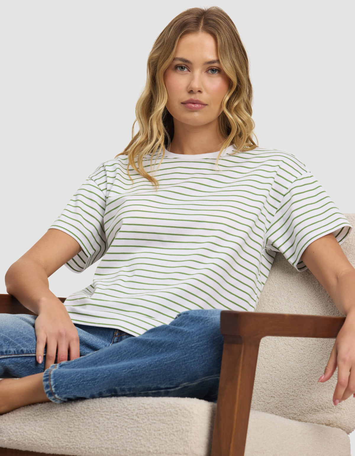 A woman with wavy blonde hair sits on a light-colored armchair, wearing Cozy Earth's Women's All Day Tee and blue jeans, looking directly at the camera with a neutral expression.|Color:Clover Stripe