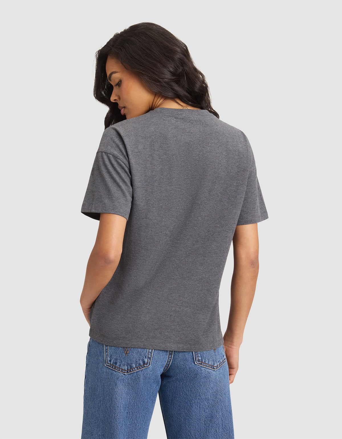 A woman with long dark hair, wearing the Cozy Earth Women's All Day Tee in gray and blue jeans, stands facing away with hands in her pockets against a plain light background. |Color:Coal Heather