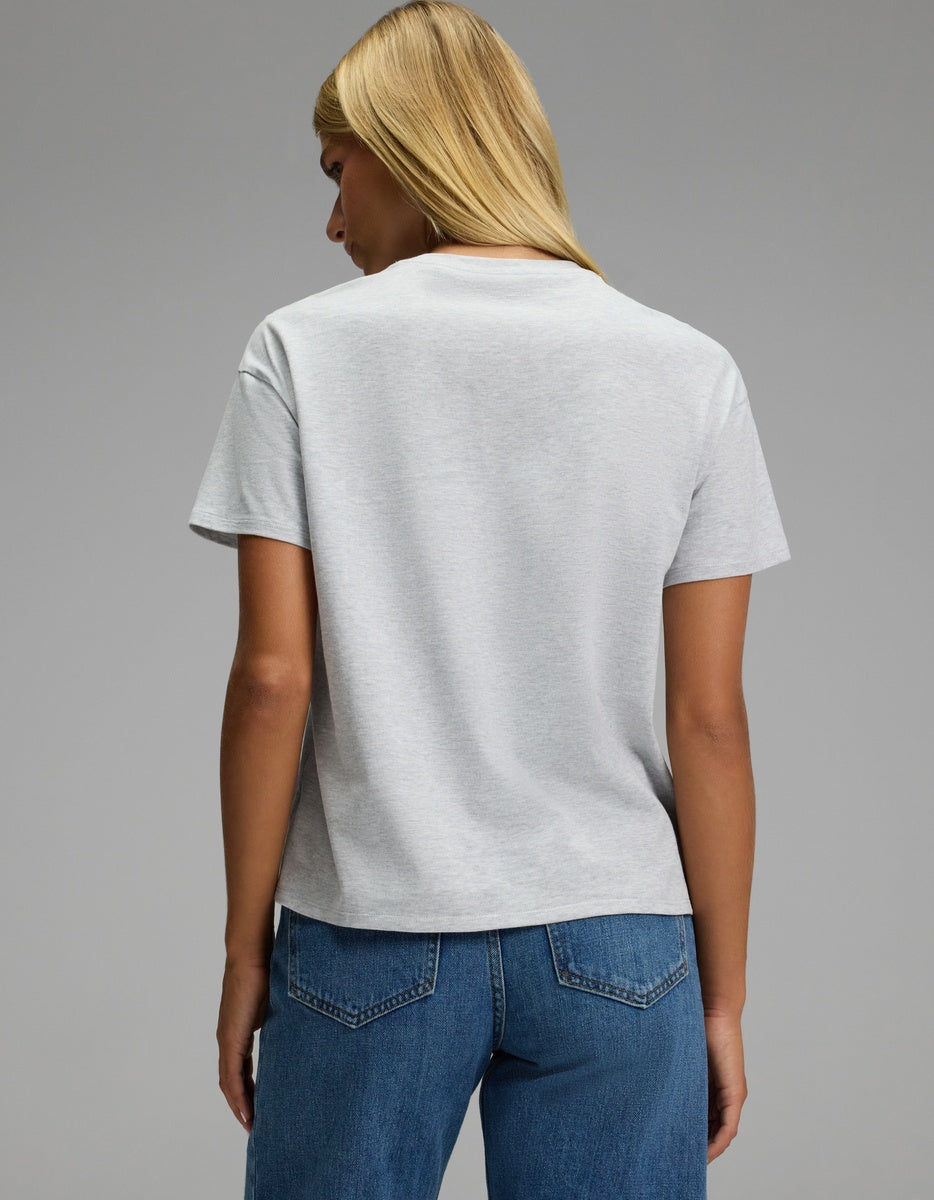 A person with straight blonde hair wears the Cozy Earth Women’s All Day Tee in light gray, paired with blue jeans, standing with their back to the camera against a plain gray background.|Color:Dove Grey Heather