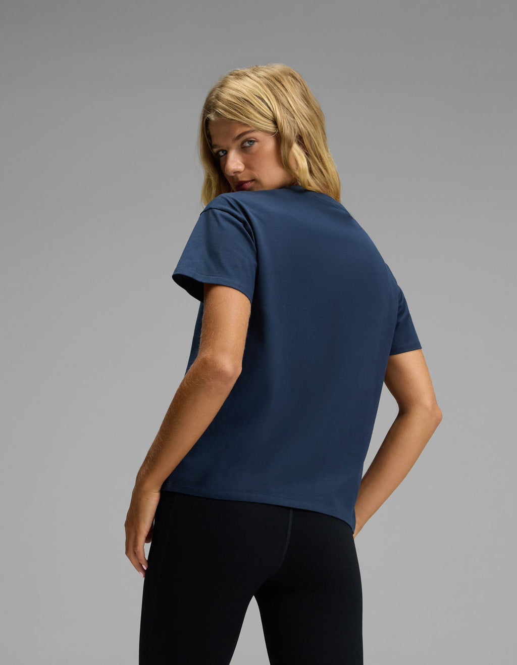 A woman with blonde hair stands with her back to the camera, turning her head to look over her shoulder while wearing Cozy Earth’s Women’s All Day Tee in navy blue and black pants, set against a plain gray background.|Color:Eclipse