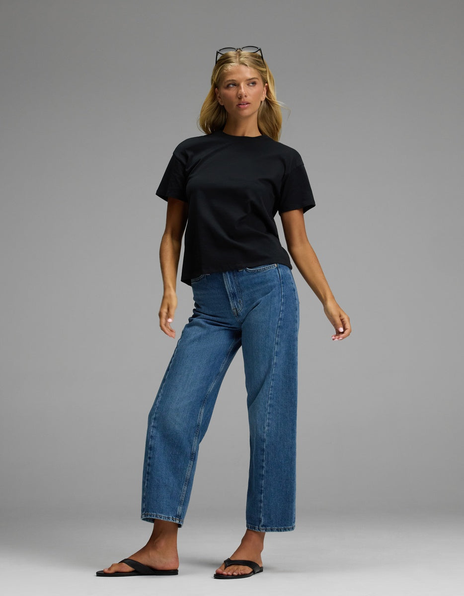 A woman models the Cozy Earth Women’s All Day Tee in black, paired with blue jeans and black sandals against a simple gray background. With blonde hair and sunglasses resting on her head, she gazes slightly to the side with a neutral expression.|Color:Jet Black