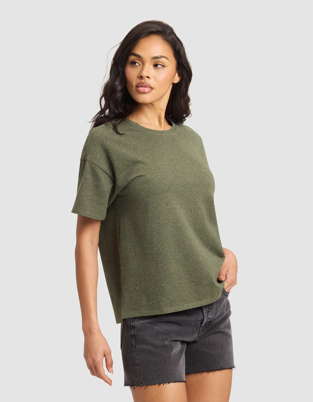 A woman with long dark hair models the Cozy Earth Women's All Day Tee in olive green, paired with black denim shorts. She stands against a plain light gray background, looking slightly left with one hand in her pocket.|Color:Olive Heather