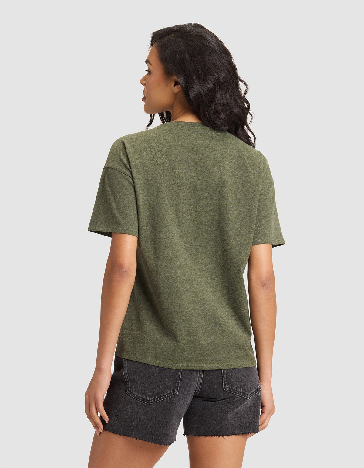 A woman with wavy dark hair stands facing away, wearing the Cozy Earth Women's All Day Tee in green and black denim shorts, set against a plain light gray background. |Color:Olive Heather