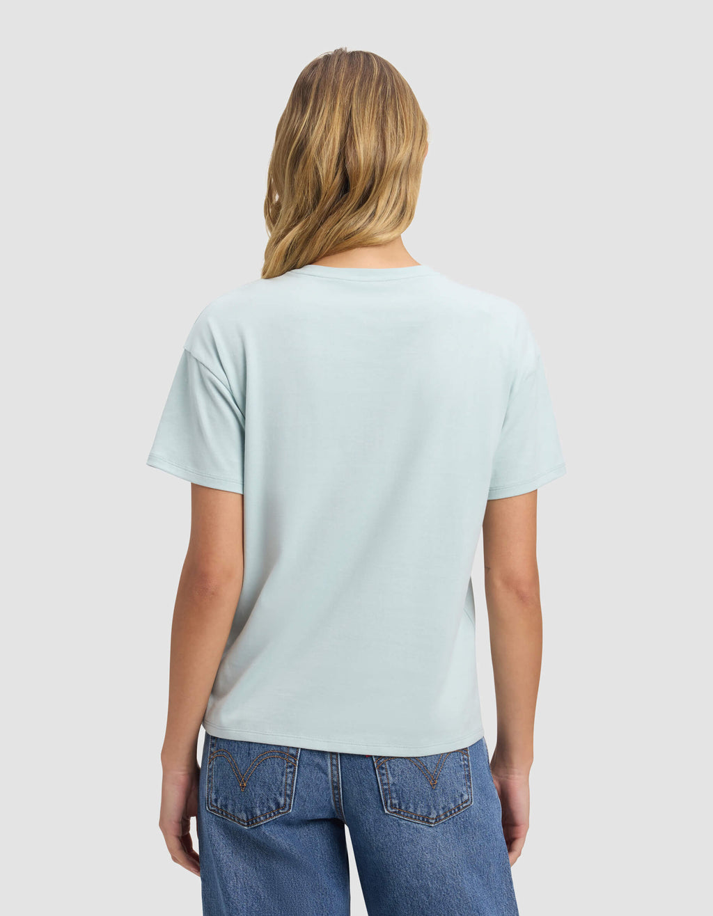 A woman with long blonde hair, dressed in Cozy Earth's Women's All Day Tee in light blue and blue jeans, faces away from the camera against a plain light gray background.|Color:Tide