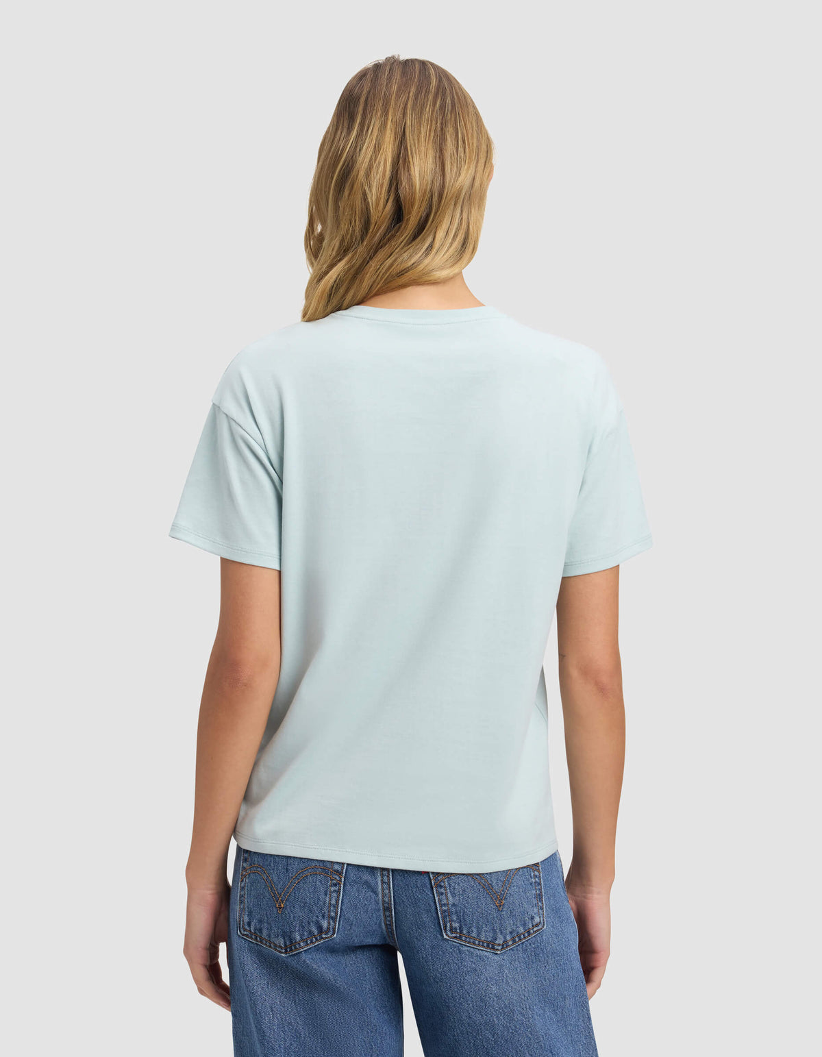 A woman with long blonde hair, dressed in Cozy Earth's Women's All Day Tee in light blue and blue jeans, faces away from the camera against a plain light gray background.|Color:Tide
