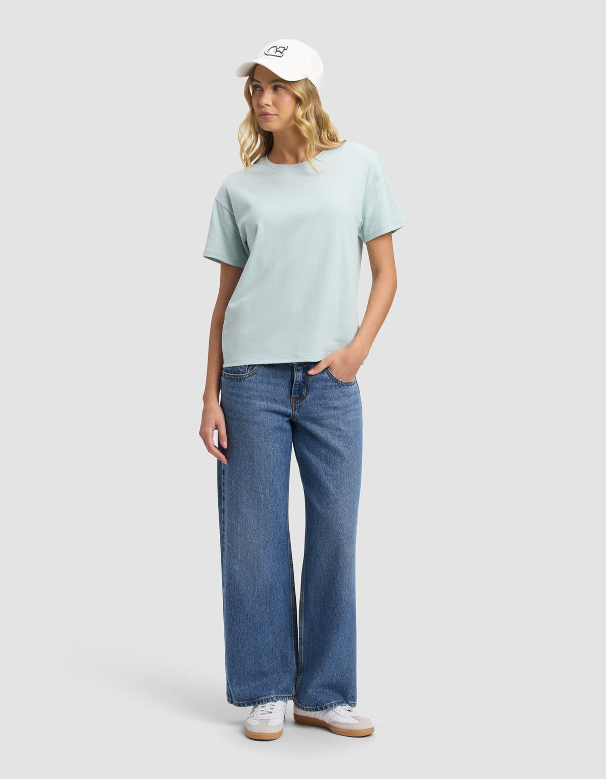 A person stands against a plain light gray background, wearing Cozy Earth’s Women's All Day Tee in light blue, blue wide-leg jeans, white sneakers, and a white baseball cap, looking slightly to the side with one hand in their pocket.|Color:Tide