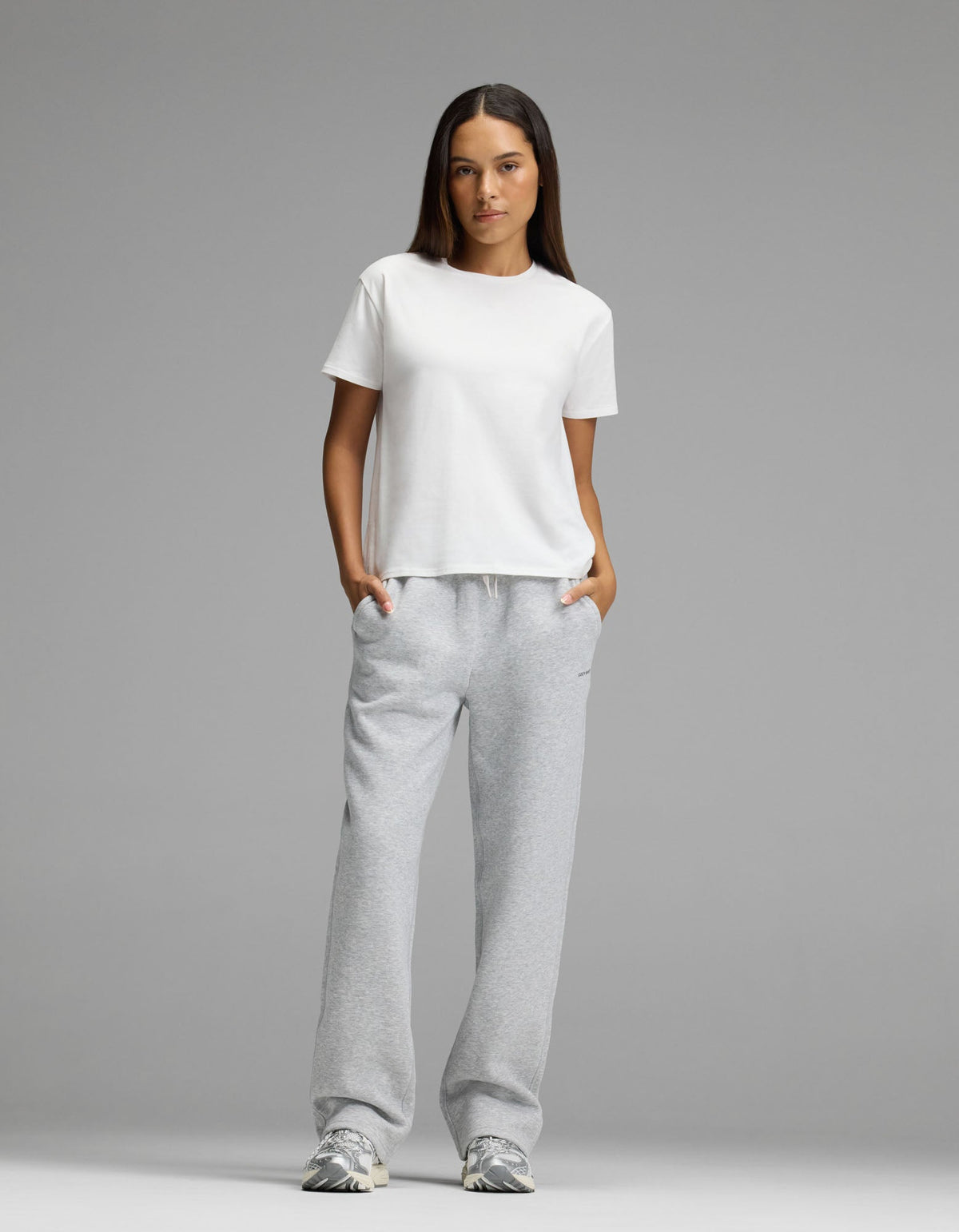 A woman stands facing forward against a plain gray background, wearing the Cozy Earth Women’s All Day Tee in white, light gray sweatpants, and sneakers, with her hands in her pockets.|Color:White