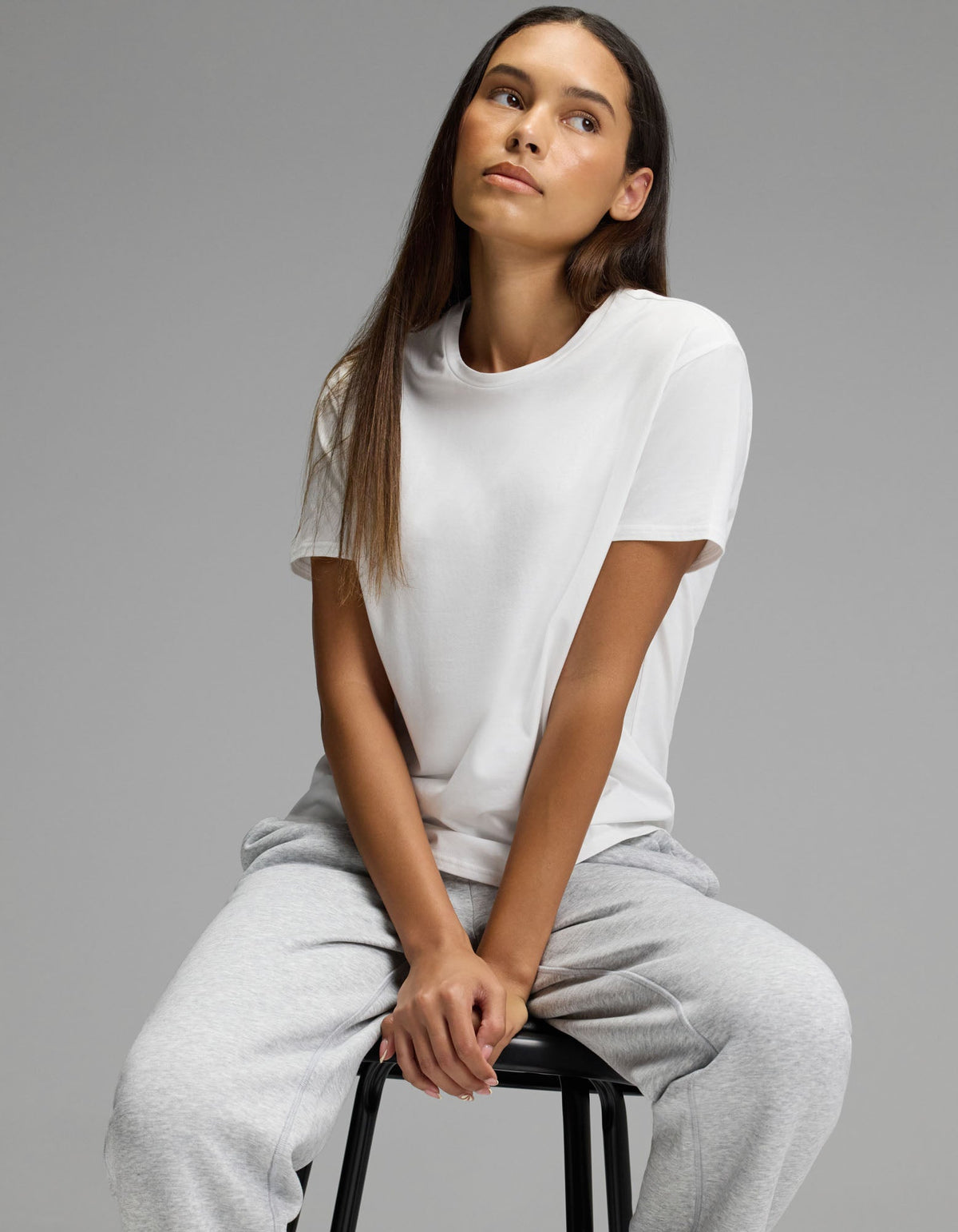 A woman wearing the Cozy Earth Women’s All Day Tee in white and gray sweatpants sits on a black stool against a plain gray background, looking slightly up and to the side with a relaxed expression.|Color:White