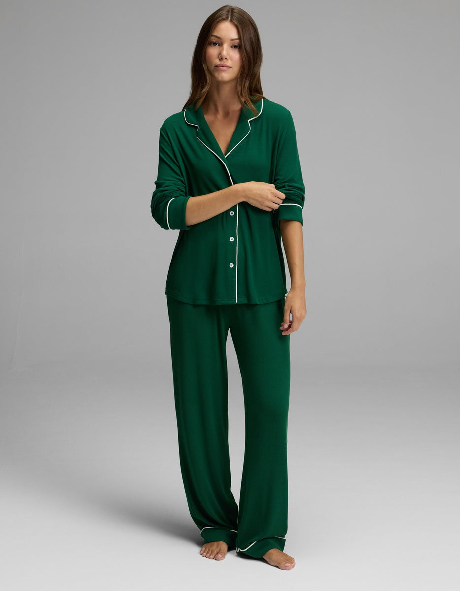 A woman stands barefoot against a plain light gray background, wearing the Cozy Earth Women’s Bamboo Rib-Knit Classic Long Sleeve Pajama Top in dark green with white piping, paired with matching loose pants.|Color:Evergreen