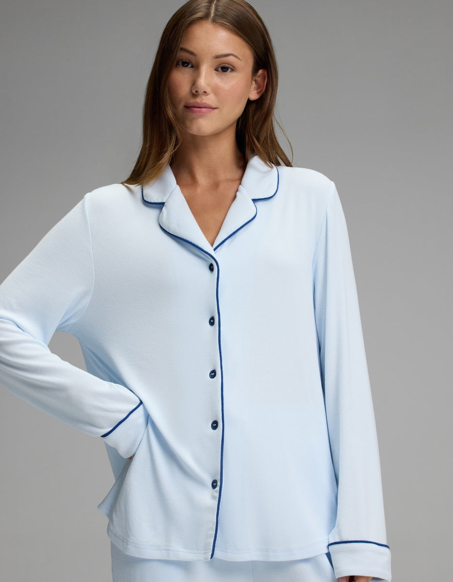 A woman models the Cozy Earth Women’s Bamboo Rib-Knit Classic Long Sleeve Pajama Top in light blue with dark trim, standing with one hand on her hip and looking at the camera against a plain gray background.|Color:Frost