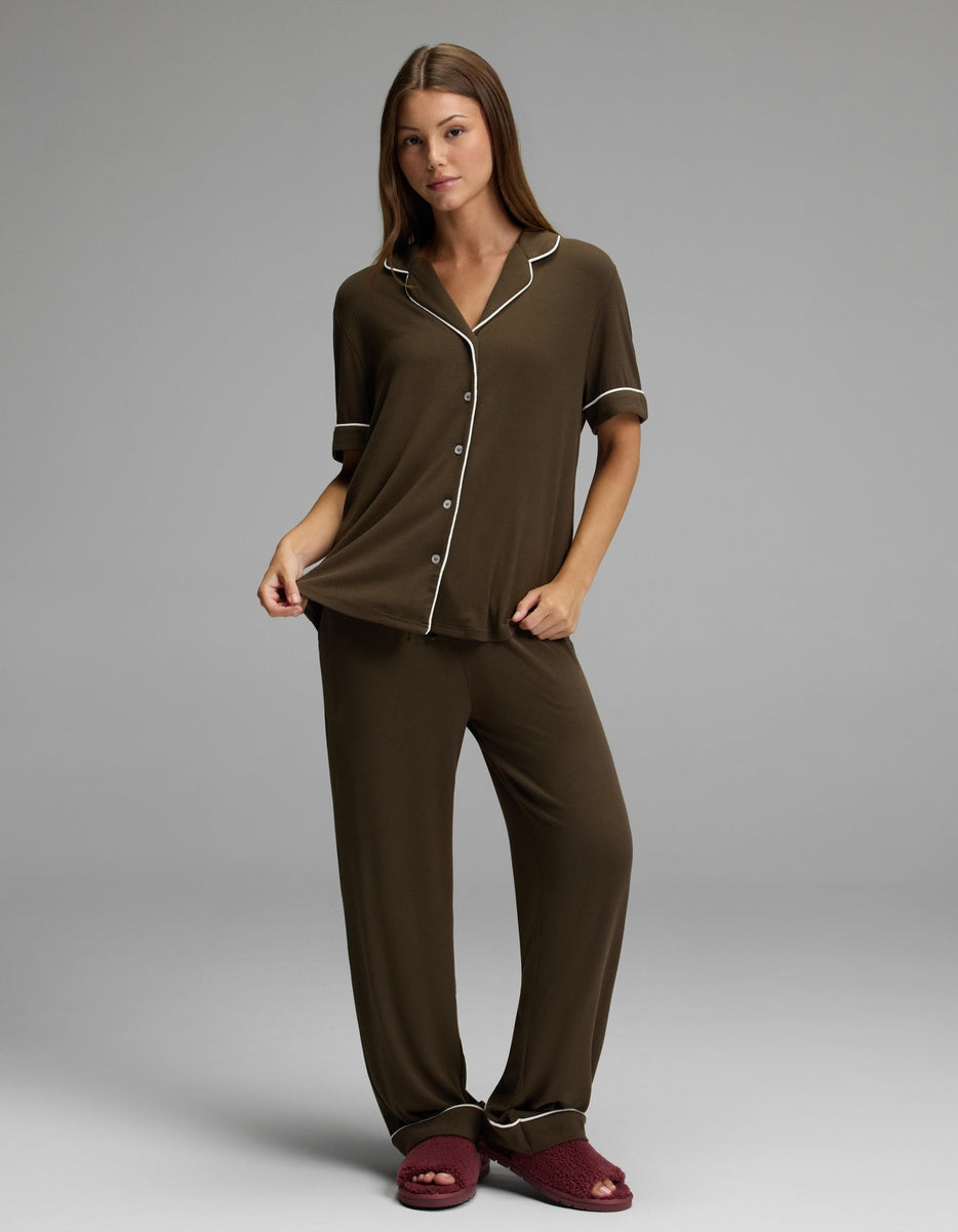 A woman stands against a plain grey background, holding the edge of her Cozy Earth Women’s Bamboo Rib-Knit Classic Short Sleeve Pajama Top in brown with white piping, and wears red slippers.|Color:Espresso