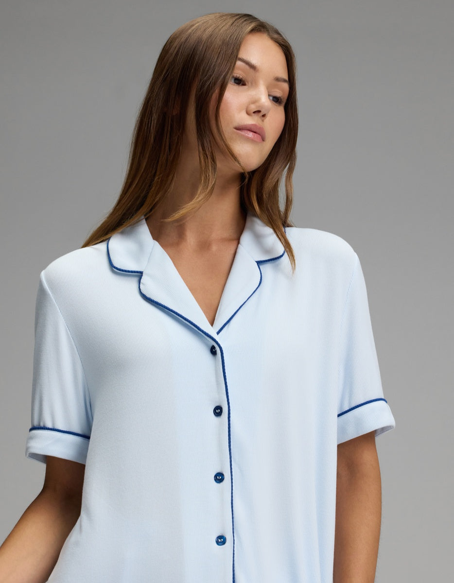 A woman with long brown hair wears the Cozy Earth Women’s Bamboo Rib-Knit Classic Short Sleeve Pajama Top in light blue, featuring dark piping, as she poses against a plain gray background.|Color:Frost