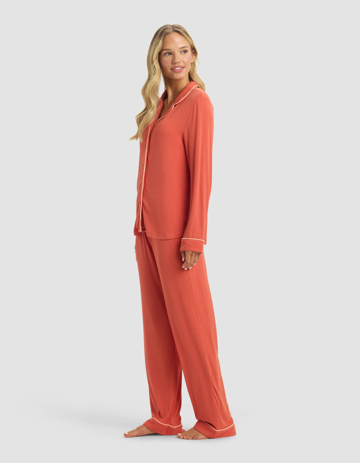 A woman with long, wavy blonde hair smiles gently as she stands barefoot, angled to the side, wearing Cozy Earth's Women's Bamboo Rib-Knit Classic Long Sleeve Pajama Set in coral with white piping, featuring a button-down top and loose pants.|Color:Sunset
