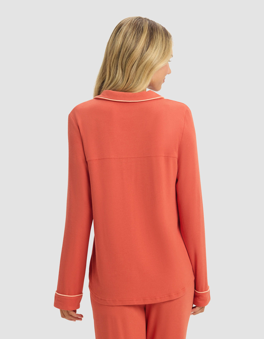 A woman with blonde hair is seen from behind, wearing a coral Women's Bamboo Rib-Knit Classic Long Sleeve Pajama Top by Cozy Earth, standing against a plain light gray background.|Color:Sunset