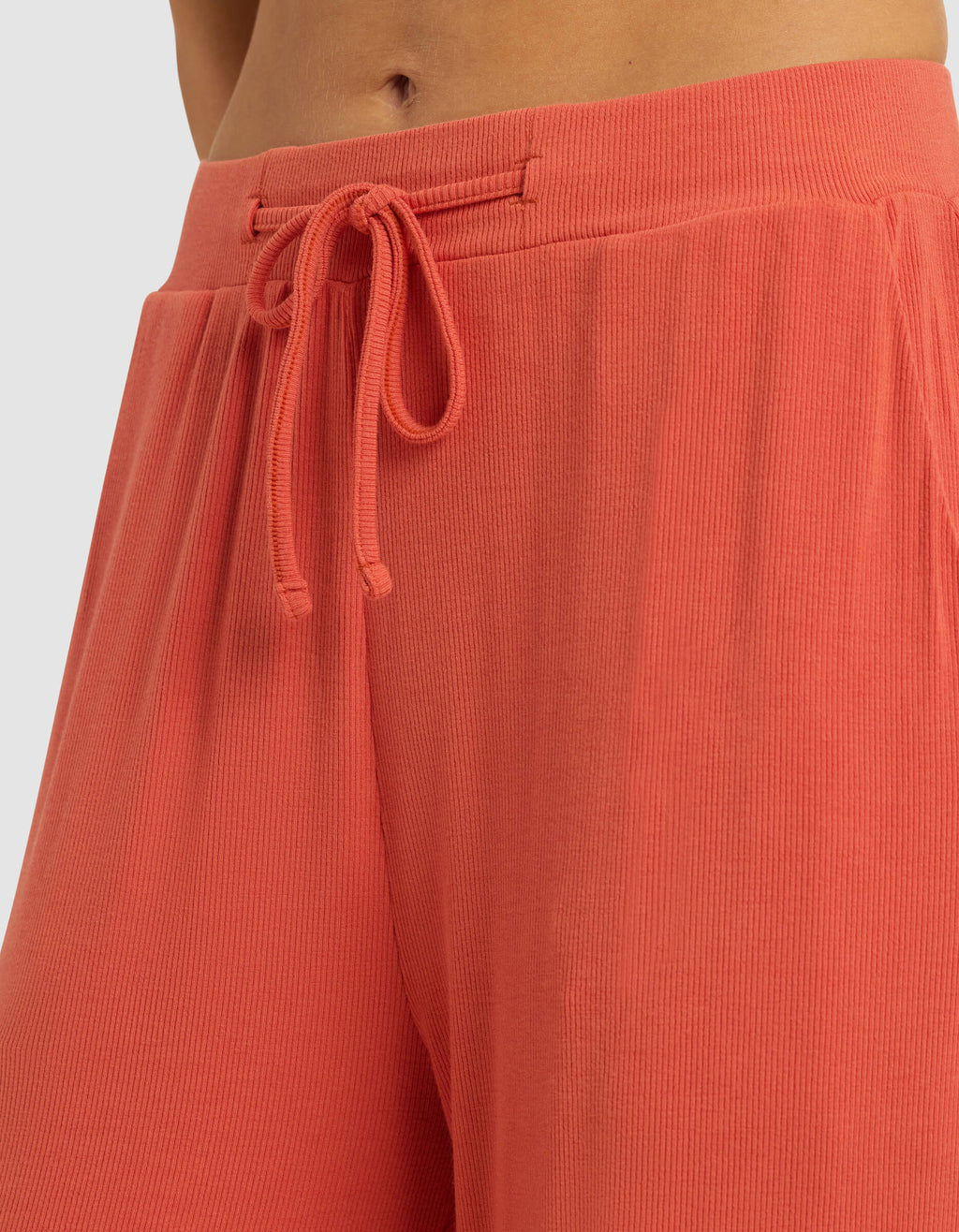 Close-up of a person wearing Cozy Earth Women's Bamboo Rib-Knit Classic Pajama Pant in coral, featuring a high waist, ribbed texture, and drawstring tie. Only the lower torso and upper legs are shown; fabric looks soft and comfortable.|Color:Sunset