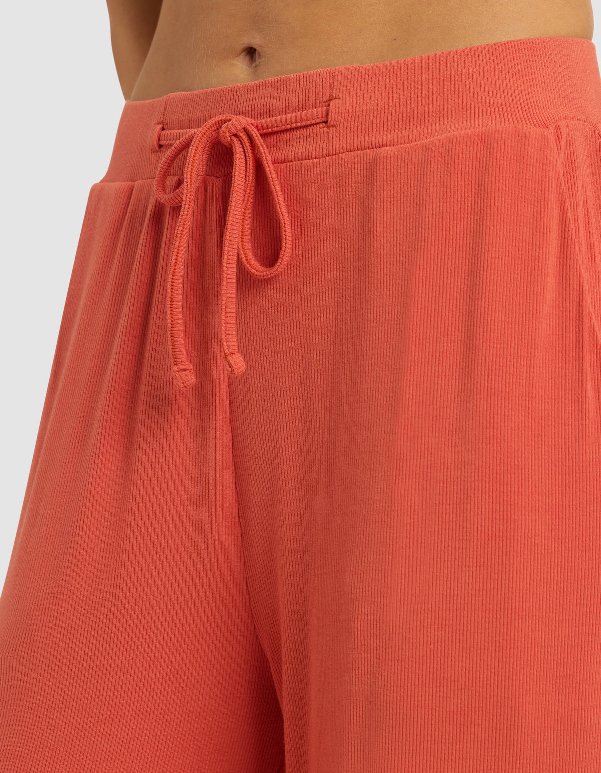 Close-up of a person wearing Cozy Earth Women's Bamboo Rib-Knit Classic Pajama Pant in coral, featuring a high waist, ribbed texture, and drawstring tie. Only the lower torso and upper legs are shown; fabric looks soft and comfortable.|Color:Sunset