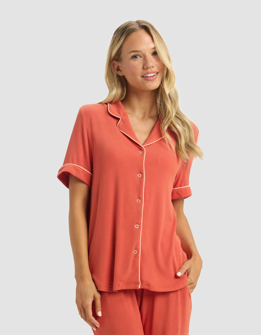 A woman with long blonde hair smiles in the Cozy Earth Women's Bamboo Rib-Knit Classic Short Sleeve Pajama Set, a coral ensemble with white piping, button-up top, and matching pants, standing against a plain light background.|Color:Sunset