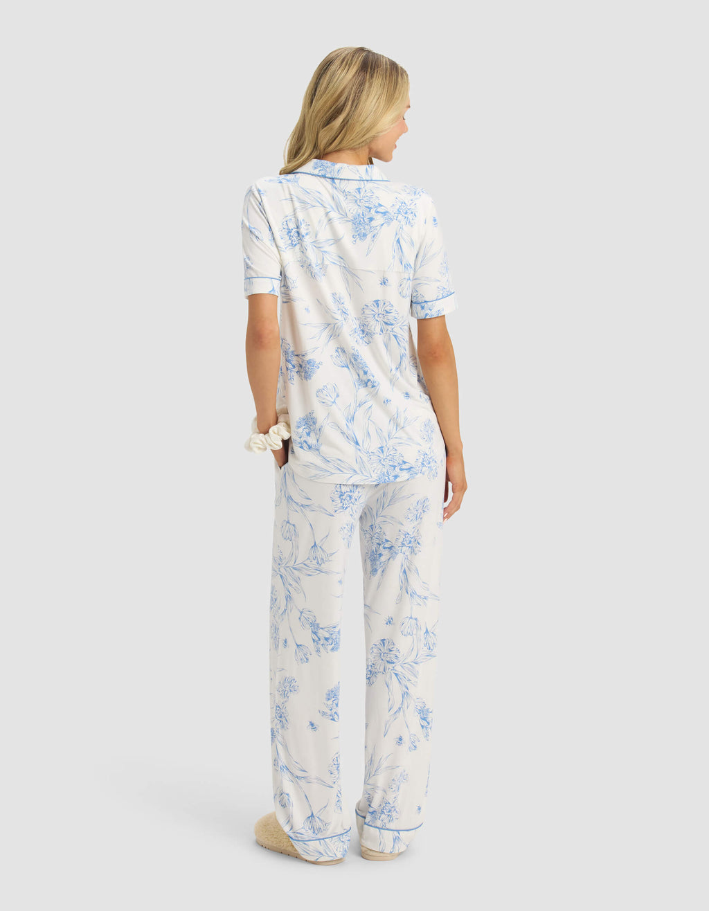 A woman with blonde hair faces away, wearing the Cozy Earth Women's Bamboo Stretch-Knit Short Sleeve & Pant Pajama Set in white with light blue floral print. She has a scrunchie on her wrist and is wearing fuzzy slippers.|Color:Garden Toile in Sea Salt