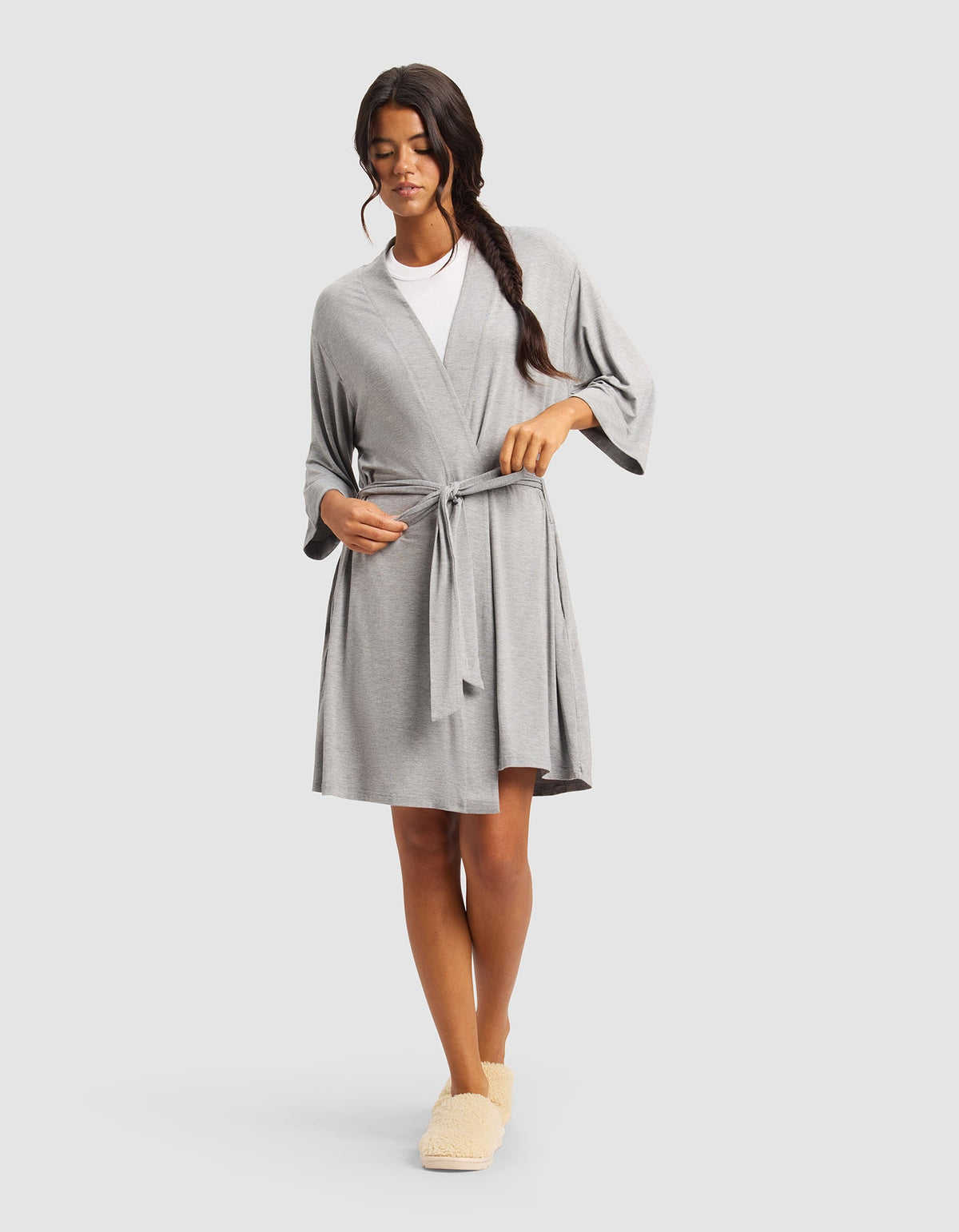 A woman with long dark hair in a side braid wears the Cozy Earth Women's Bamboo Stretch-Knit Kimono Robe, tied at the waist over a white top and beige slippers, standing against a plain light gray background.|Color:Grey