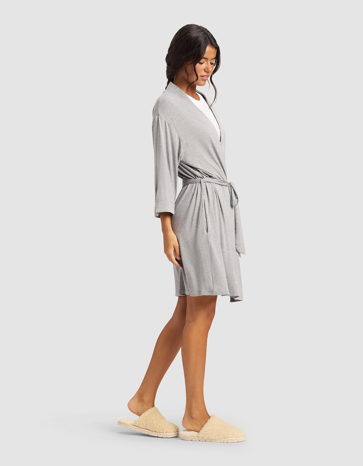 A woman stands sideways against a plain light background, relaxed in Cozy Earth’s Women's Bamboo Stretch-Knit Kimono Robe in light gray with a waist tie, paired with beige slippers. Her loose waves add to the comfortable vibe.|Color:Grey