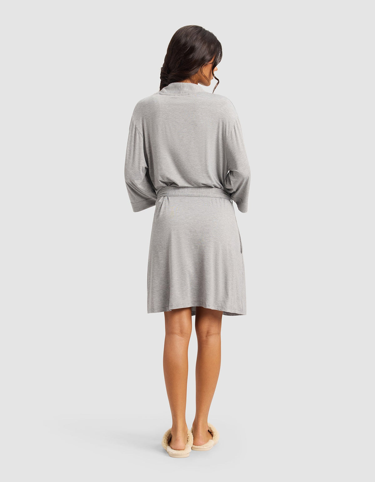 A woman with long dark hair, seen from the back, wears a Cozy Earth Women's Bamboo Stretch-Knit Kimono Robe in light gray with a belt and cream slippers, standing against a plain light background.|Color:Grey