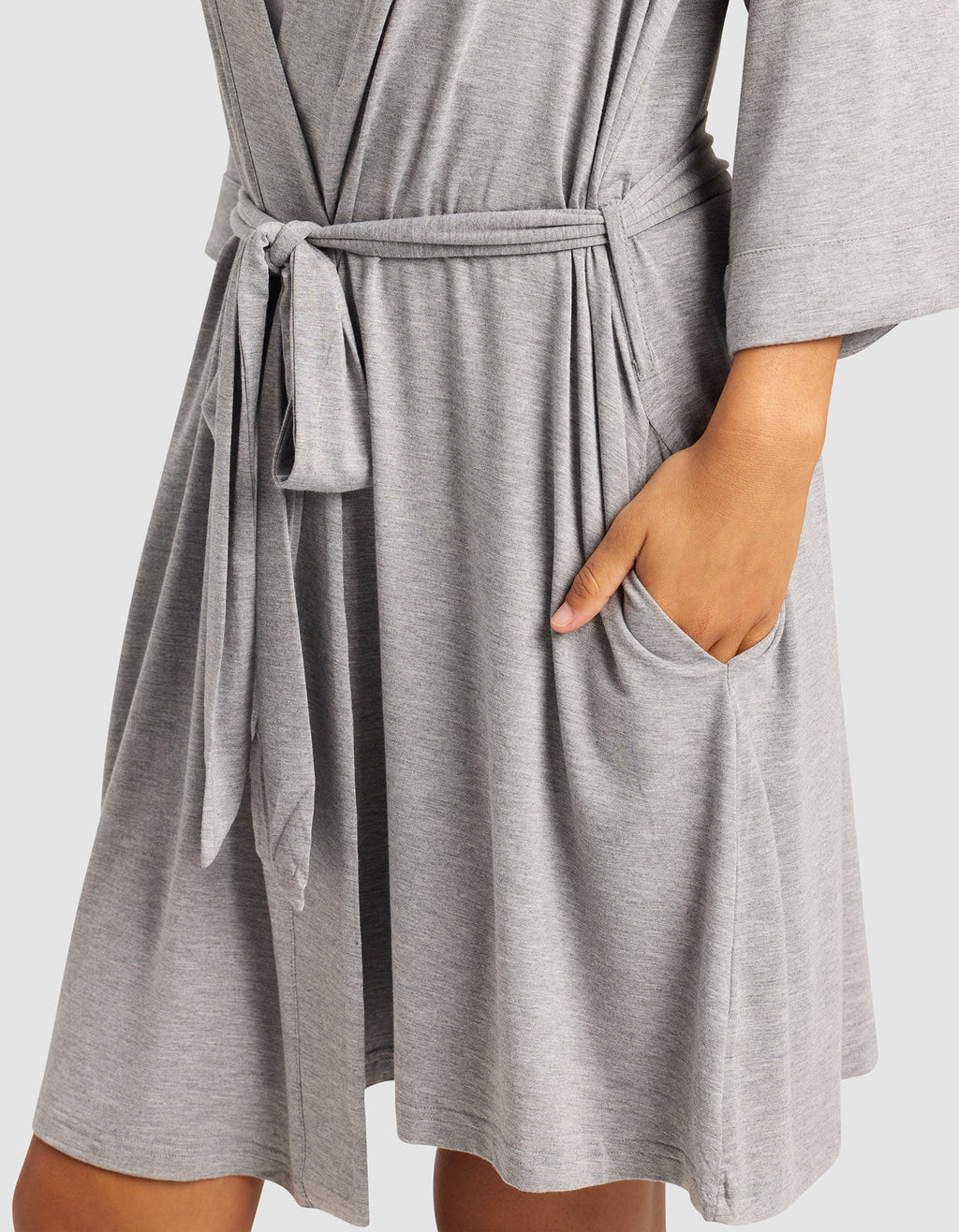 A person wears Cozy Earth's Women's Bamboo Stretch-Knit Kimono Robe in light gray, featuring long sleeves, a relaxed fit, knee length, fabric belt at the waist, and pockets with their right hand tucked in.|Color:Grey