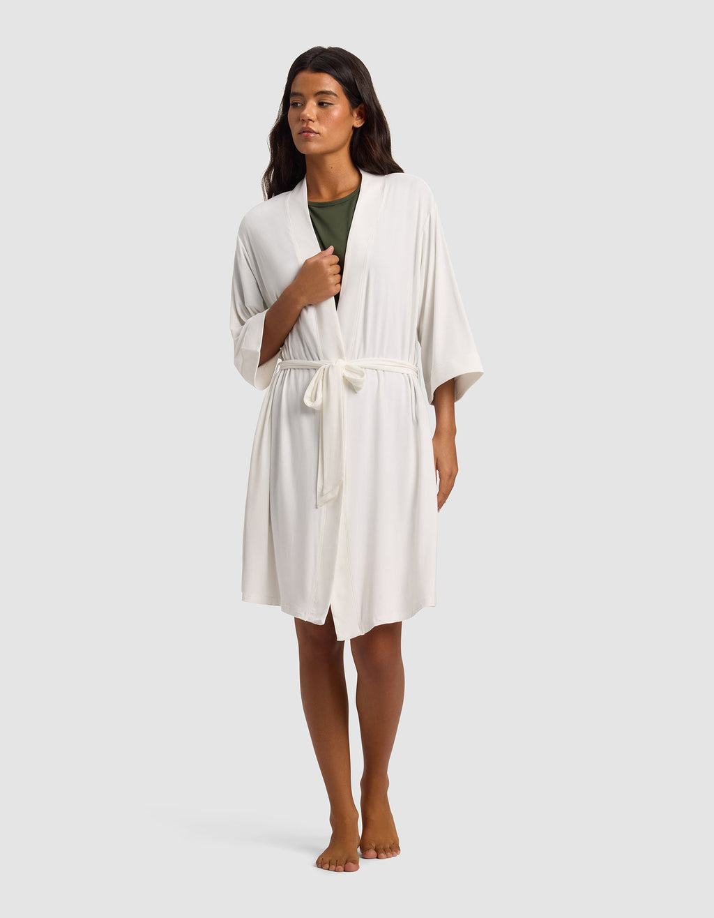 A person stands barefoot against a plain light gray background, wearing Cozy Earth's Women's Bamboo Stretch-Knit Kimono Robe in white with a tie belt over a green top.|Color:Ivory