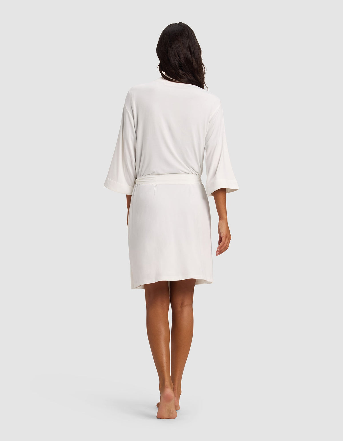 A woman with long dark hair stands barefoot, facing away, wearing the Cozy Earth Women's Bamboo Stretch-Knit Kimono Robe—a knee-length white robe with loose sleeves and a belt—against a plain light gray background.|Color:Ivory