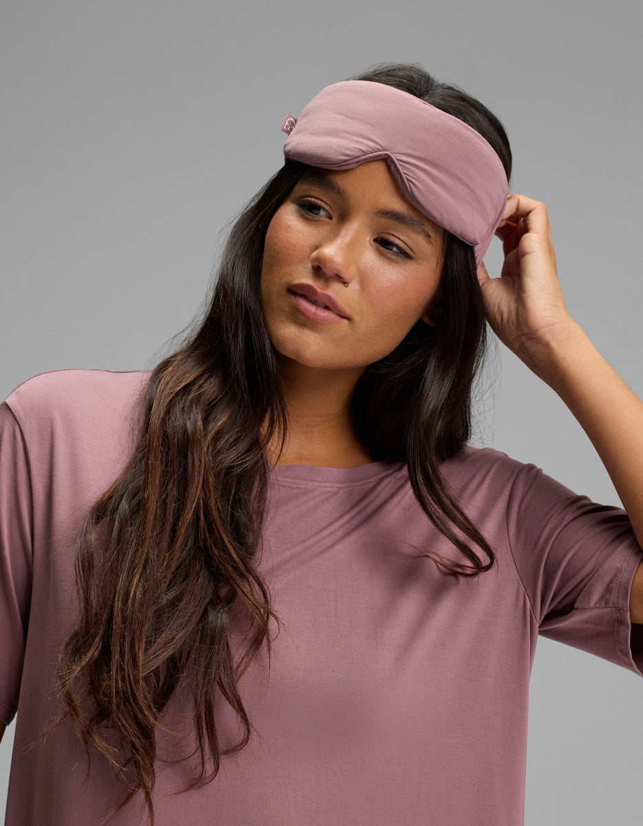 A woman wearing a mauve Bamboo Stretch-Knit Sleep Mask on her forehead and a matching short-sleeved top.|Color:Copper Rose