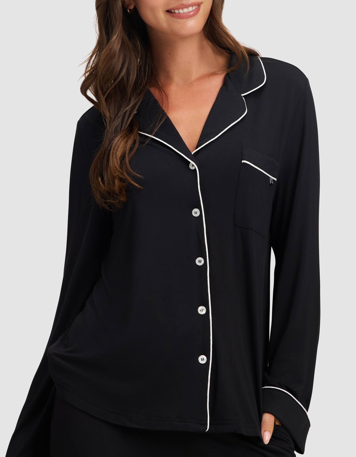 A woman smiles with one hand in her pocket, wearing the Cozy Earth Women's Bamboo Stretch-Knit Long Sleeve Pajama Top in black with white piping, white buttons, and a chest pocket against a plain background. |Color:Black