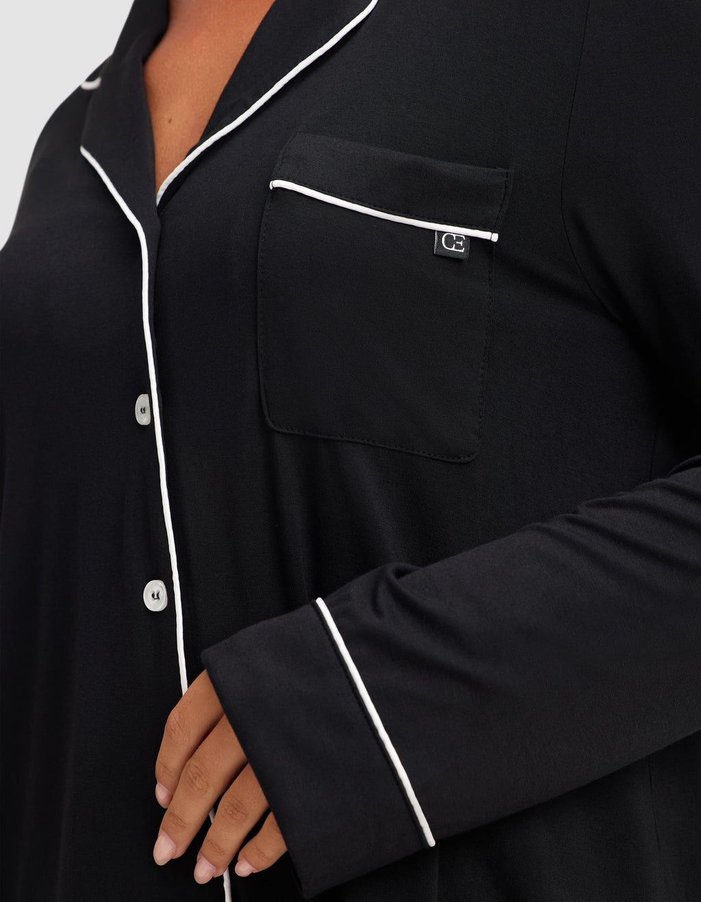 A close-up shows a person wearing the HIDE Women's Bamboo Stretch-Knit Long Sleeve Pajama Top in black, featuring white piping on the collar, pocket, and sleeve, with four white buttons visible and a hand resting near the waist. |Color:Black