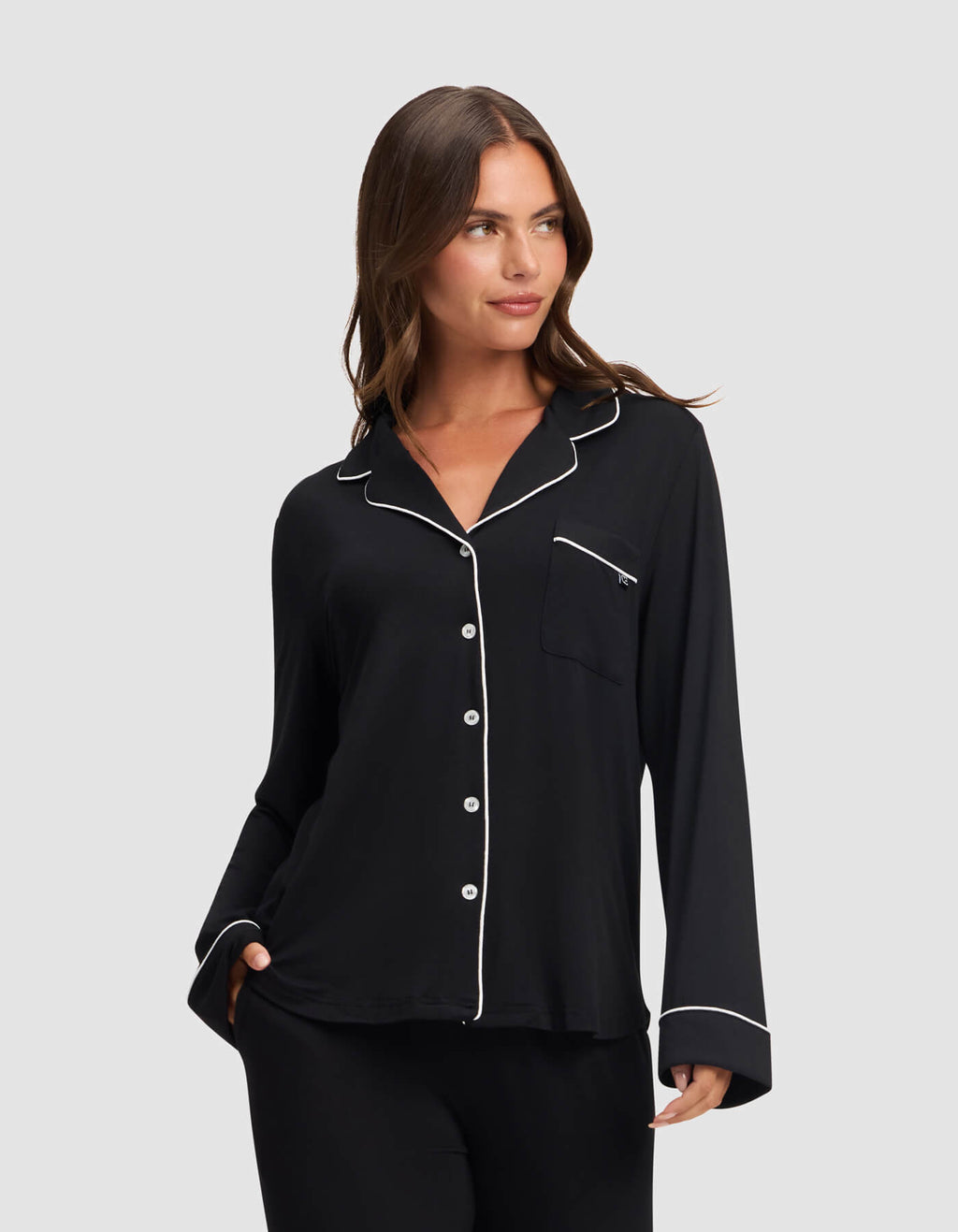 A woman models the Cozy Earth Women's Bamboo Stretch-Knit Long Sleeve Pajama Top in black with white piping on the collar, cuffs, and pocket edges, standing against a light gray background and smiling softly. |Color:Black