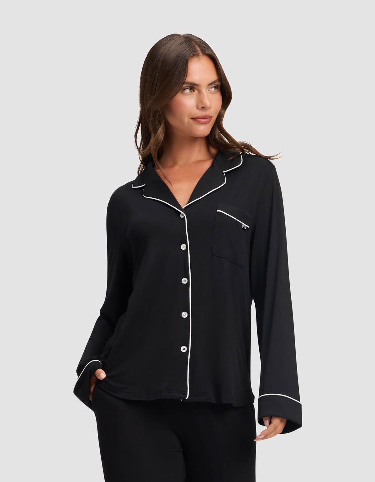 A woman models the Cozy Earth Women's Bamboo Stretch-Knit Long Sleeve Pajama Top in black with white piping on the collar, cuffs, and pocket edges, standing against a light gray background and smiling softly. |Color:Black
