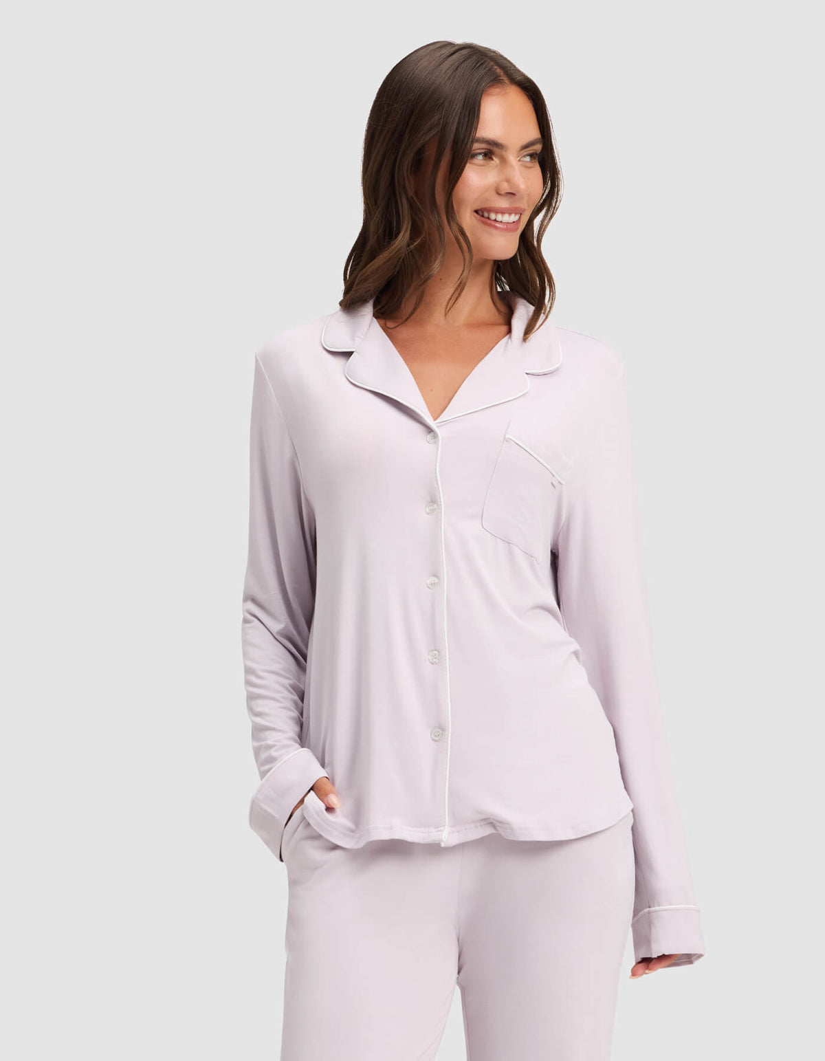 A woman smiles while wearing the Cozy Earth Women's Bamboo Stretch-Knit Long Sleeve Pajama Top in light lavender with a chest pocket, paired with matching pants. She has one hand in her pocket and stands against a plain light gray background.|Color:Lilac