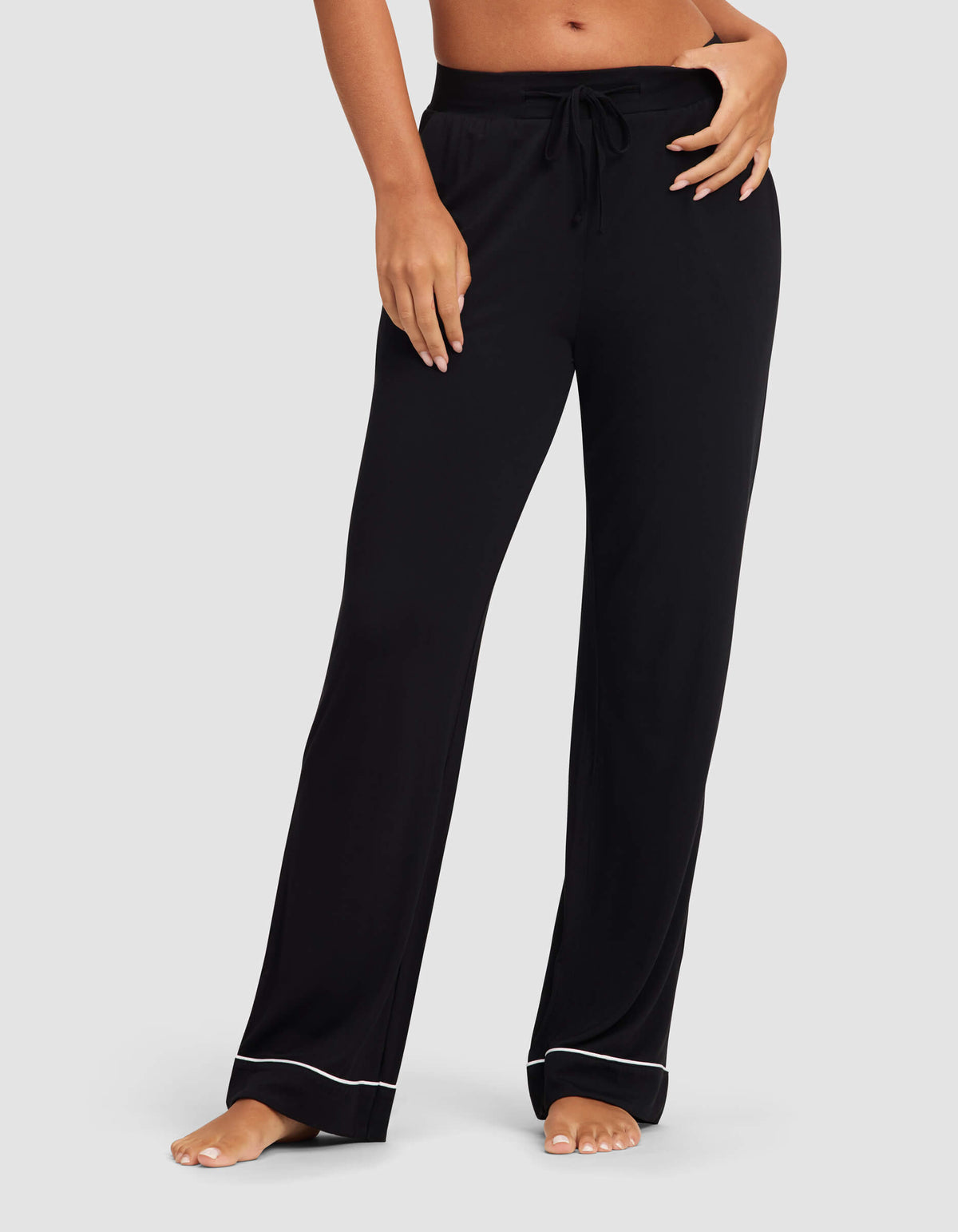 A woman stands barefoot against a plain background wearing Cozy Earth's Women's Bamboo Stretch-Knit Pajama Pant, featuring a loose fit, drawstring waist, and white piping near the hems.|Color:Black