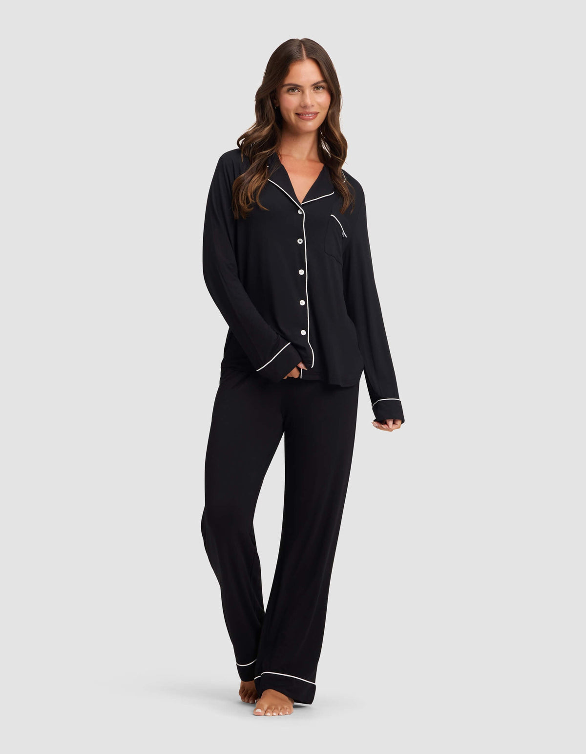 A woman with long brown hair wears Cozy Earth's Women's Bamboo Stretch-Knit Pajama Pant in black with white piping and buttons, standing barefoot and smiling against a plain light gray background.|Color:Black